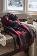 A Luxury Square Soft Red and Black Plaid 100% Baby Alpaca Wool Scarf and a beige knit blanket are draped on a rustic wooden bench beside a steaming mug, with snowy scenery visible outside the window.