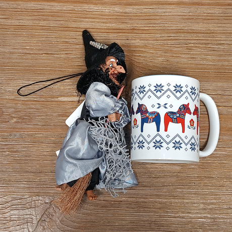 The Figurine: Kitchen Witch Doll – Sirene Silver (10.2" Tall) lies with her broom beside a white mug with colorful horses and blue patterns, displayed on a wooden surface.