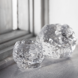 Two clear, textured KostaBoda: Snowball Votive Small 60mm candle holders by Ann Warff sit on a white surface near a window, light reflecting off their uneven surfaces.