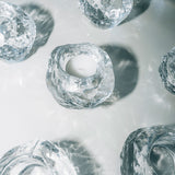 Several KostaBoda: Snowball Votive Medium 70mm candle holders with rough, irregular edges are arranged on a white surface, inspired by Ann Wolff’s design and reflecting light to cast soft shadows.