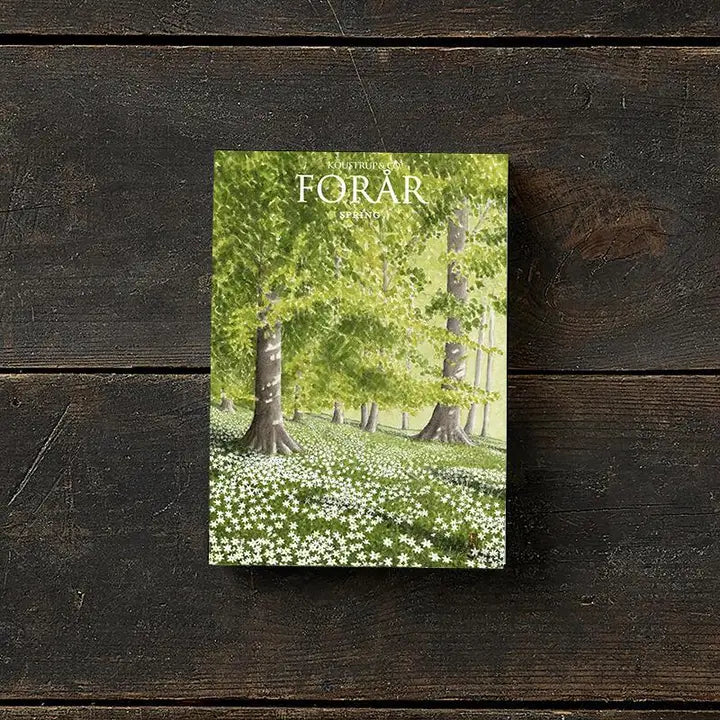 The FORÅR book with a green forest scene cover mimics the elegance of the Card Pack: Spring - 8 Doublecards w/Envelopes, featuring spring landscapes crafted from FSC paper, as it rests on a dark wooden surface.