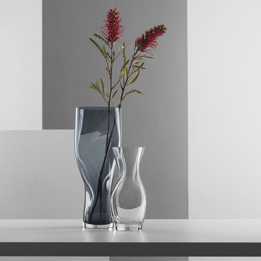 Two exquisite Orrefors vases grace the table, each showcasing elegant design. The artistic touch of Lena Bergström shines through in this harmonious display, with the Squeeze Blue/Gray Vase adding a dynamic contrast to the vibrant red flower set amidst gray and white geometric patterns.