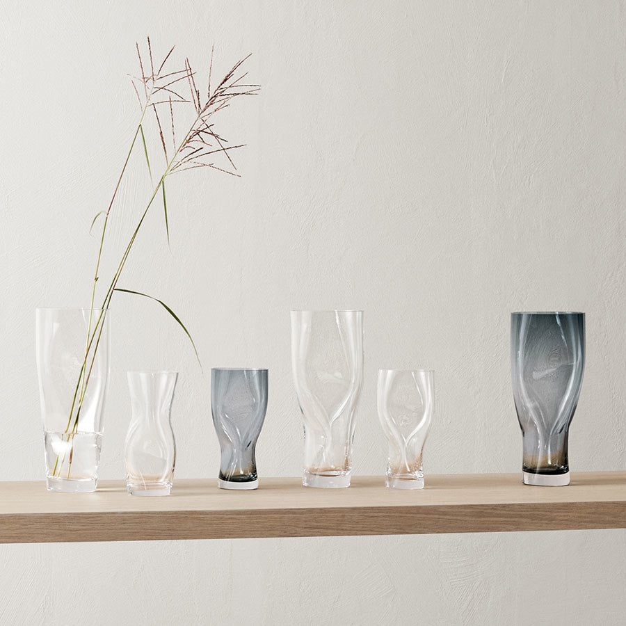 A wooden shelf displays six glass vases of different designs. The eye-catching Squeeze Blue/Gray Vase Small by Lena Bergström for Orrefors, resembling tall grass stems, stands out in the leftmost position.