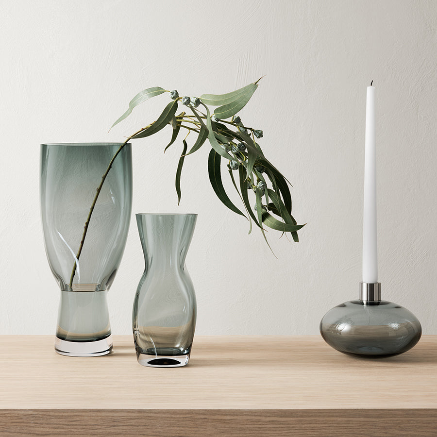 Two Orrefors Squeeze Blue/Gray vases elegantly hold eucalyptus branches beside a matching candle holder with a tall white candle on a wooden surface.