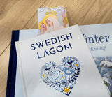 A book titled Swedish Lagom with a blue heart on the cover rests atop another, featuring a Carl Larsson “St. Bridget of Sweden” bookmark (8.5" × 2.5") that honors Scandinavian art.
