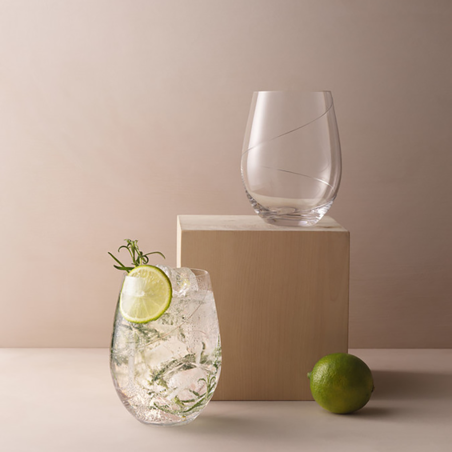 Two clear KostaBoda: Line Stemless Glass 20oz sit on a wooden block, one filled with a refreshing drink garnished with lime and herbs. A whole lime rests beside them, showcasing exquisite Swedish craftsmanship.