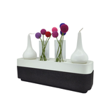 A modern vase with three glass tubes holding colorful artificial flowers stands beside two Stumpastaken Swedish Candle Holders, all displayed on a sleek black and white rectangular base.