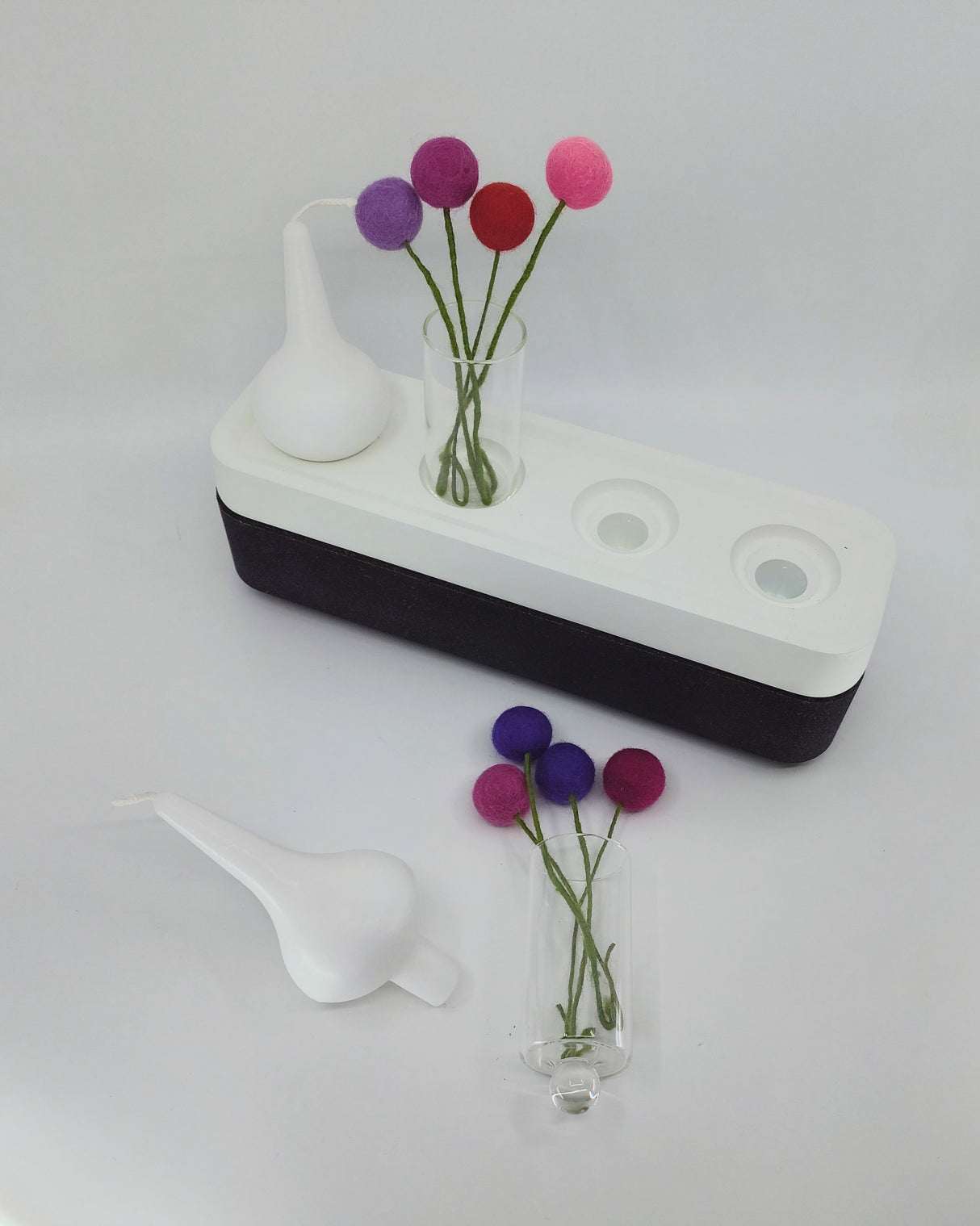 A white and black handcrafted Stumpastaken Swedish Candle Holder that holds 4 candles, with glass vases of colorful felt ball stems and a white dropper, rests on a white surface.