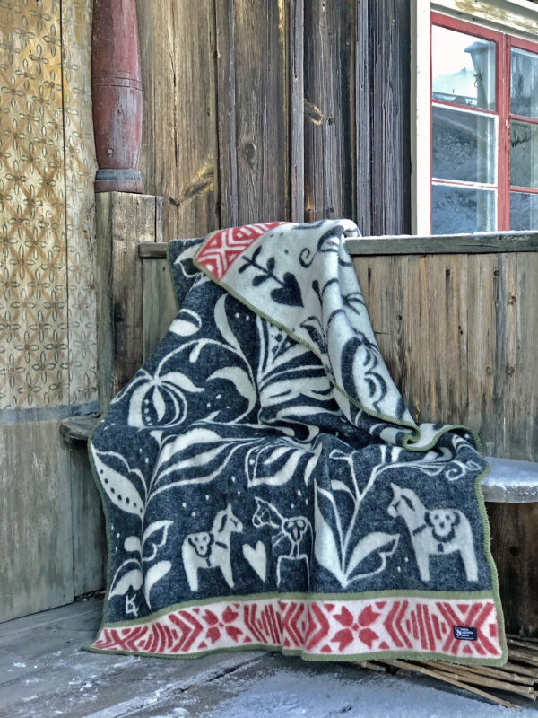 The Dalecarlia Blanket, featuring Swedish Dala Horse patterns in black, red, and white, made from 100% Norwegian Lambs Wool by Kerstin Landstrom, is draped over a rustic wooden bench against a wooden wall and window.