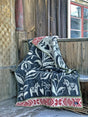 The Dalecarlia Blanket, featuring Swedish Dala Horse patterns in black, red, and white, made from 100% Norwegian Lambs Wool by Kerstin Landstrom, is draped over a rustic wooden bench against a wooden wall and window.