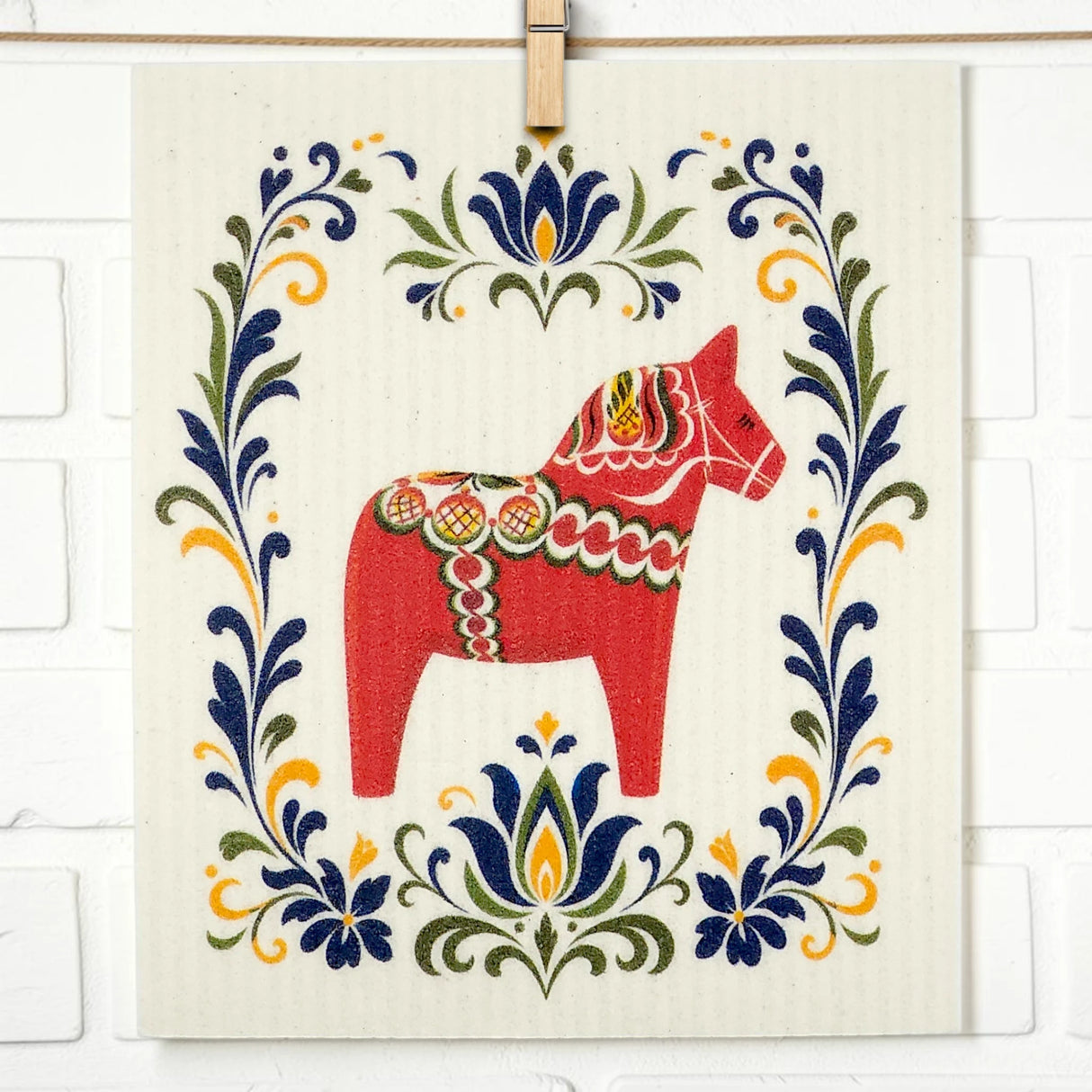 White Swedish dishcloth featuring a red Dala horse with traditional patterns, surrounded by blue, green, and yellow florals, clipped to a line against a white wall.