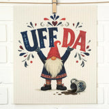 Swedish dishcloth with a gnome holding up “UFF DA,” a spilled mug and dark liquid at its feet, and decorative floral patterns in the background.