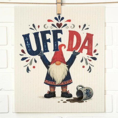 Swedish dishcloth with a gnome holding up “UFF DA,” a spilled mug and dark liquid at its feet, and decorative floral patterns in the background.