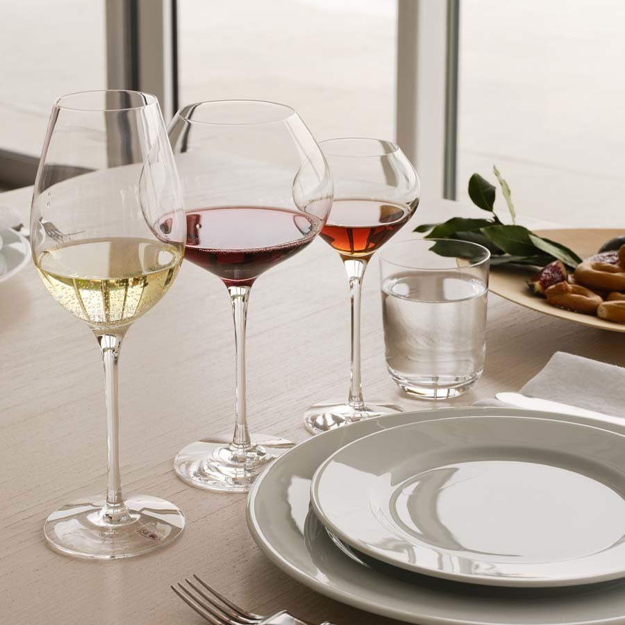 A table setting includes three wine glasses—white, red, and rosé—a water glass, and a plate. In the backdrop, figs and leaves enhance the scenes elegance. The mouth-blown Orrefors: Difference Sweet Glass 7.5oz (2-Pack) hints at an indulgence in dessert wines.