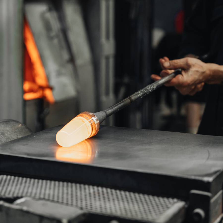 In a workshop, an artisan shapes mouth-blown molten glass with a metal rod, akin to Erika Lagerbielke’s artistry in designing the Orrefors Difference Sweet Glass 7.5oz for enjoying dessert wines.