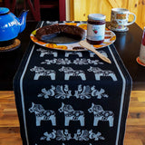 A table features a Siljan Grey runner with Swedish Dala Horses on black by Ekelund, adorned with a sliced chocolate cake on a decorative plate, a jar of lingonberry jam, and accompanied by a blue teapot and two cups.