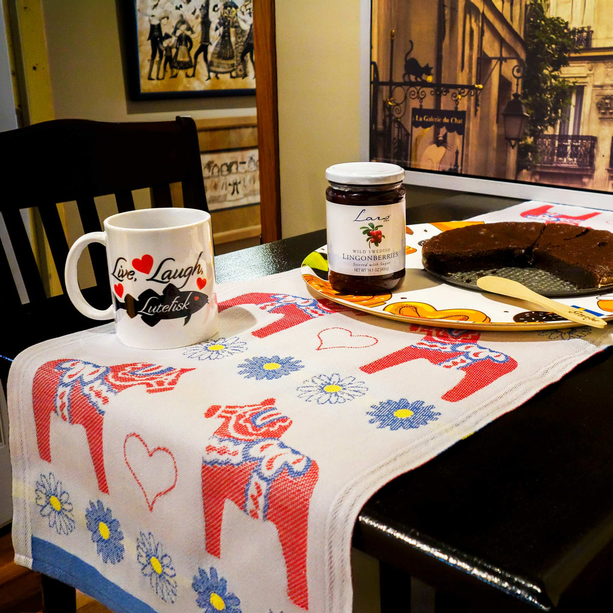A table setting includes a mug, lingonberry jam jar, and chocolate cake on a tray. The table is decorated with Ekelund Master Weavers Svea Swedish Dala Horses runner made of organic cotton, featuring bright red horses and flowers. Framed artwork and a chair are visible in the background.