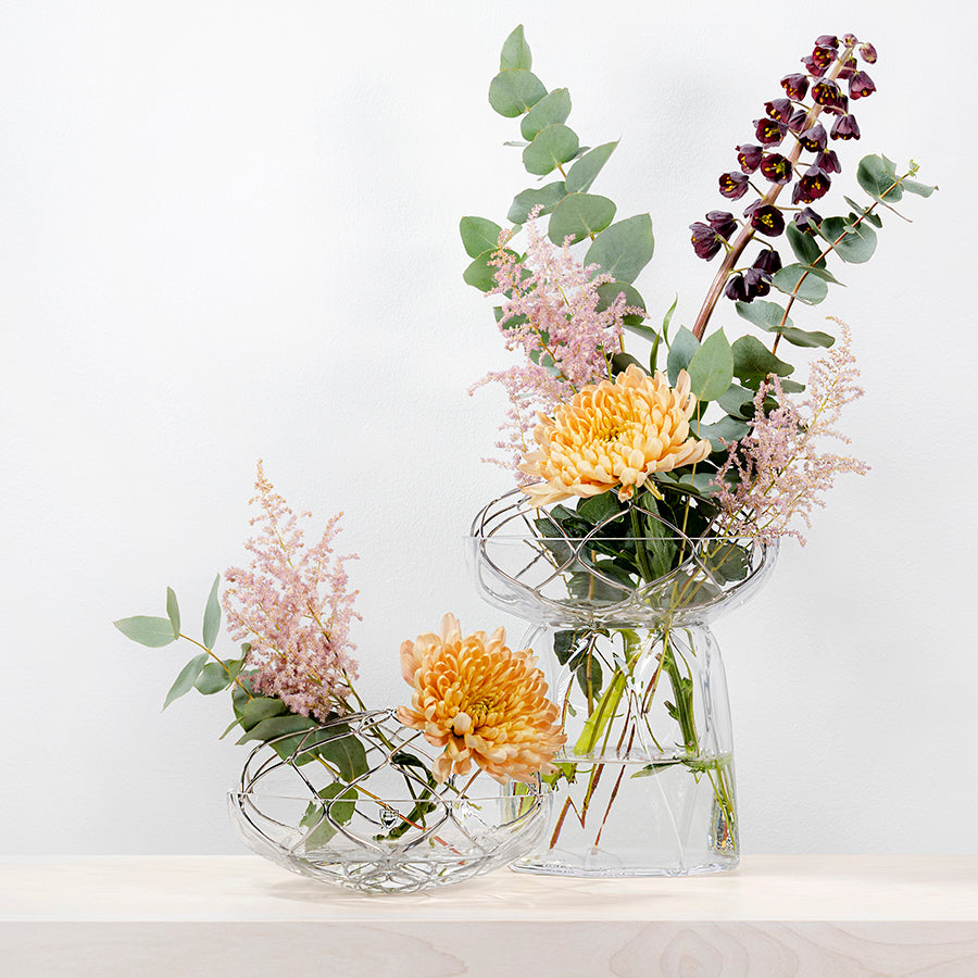 Two Orrefors: Bloom Vase Tall pieces featuring yellow and purple flowers, green leaves, and light pink accents adorn a wooden surface against a white background.