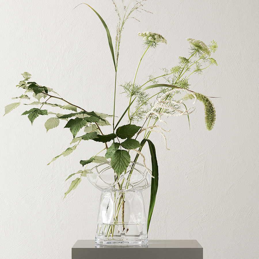 The Orrefors Bloom Vase Tall by Lisa Hilland showcases a vibrant mix of green leaves and wildflowers on a gray pedestal set against an off-white background.