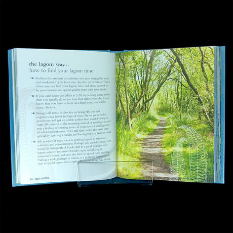 An open copy of "The Lagom Life: A Swedish Way of Living" by Elisabeth Carlsson displays tips on finding lagom time on the left page and a sunlit forest path lined with green trees on the right.