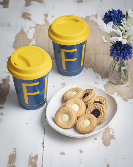 A "Fika" To-Go coffee mug with a yellow lid and blue sleeve sits next to a plate of assorted cookies and a small vase of blue and white flowers on a rustic white table.