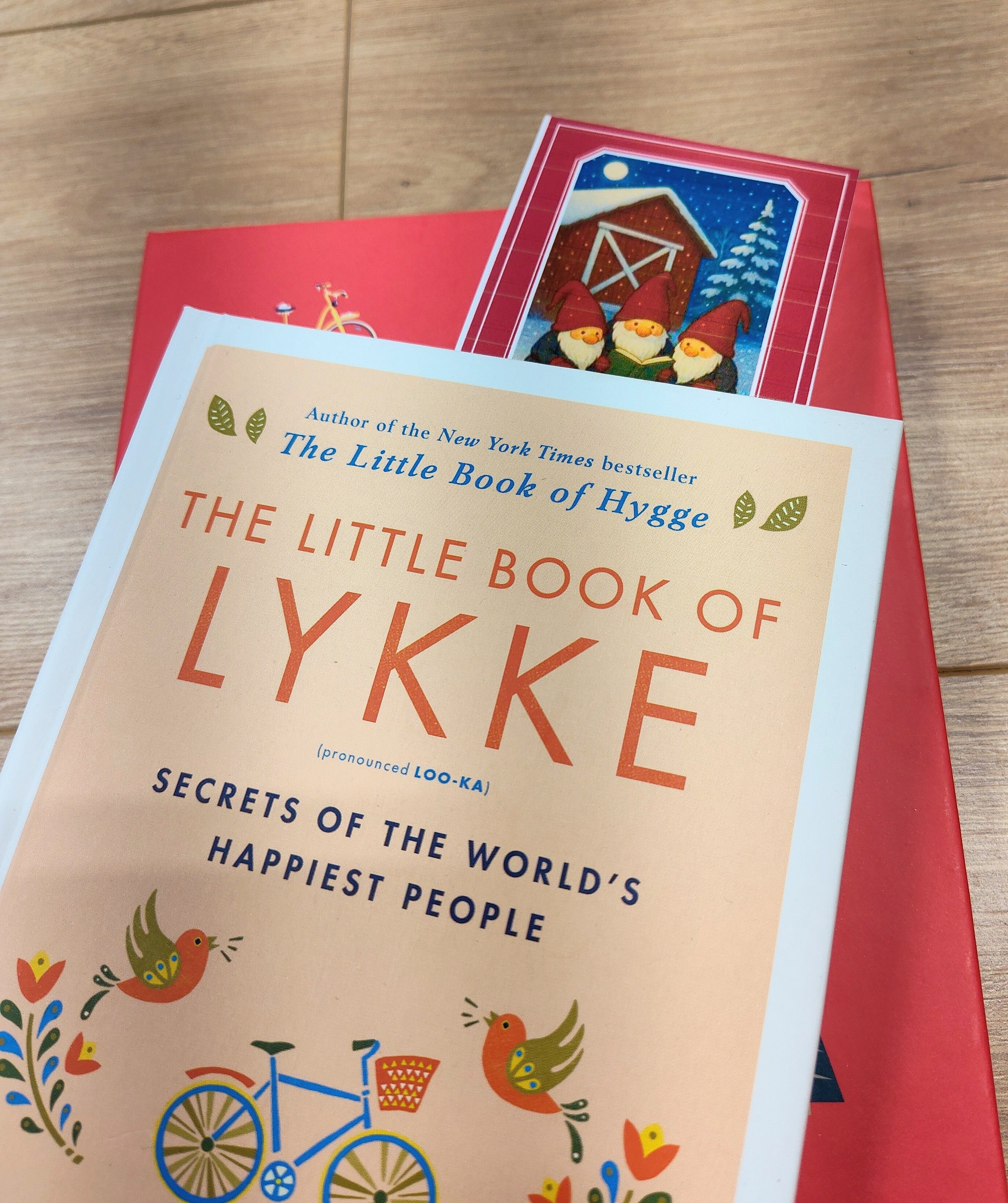 Two books rest on a wooden surface: “The Little Book of Lykke” atop a red, folklore-inspired volume with a barn and birds. A “Tomte Rules” bookmark (2.5" × 8.5") peeks out, lending Nordic winter charm to the scene.