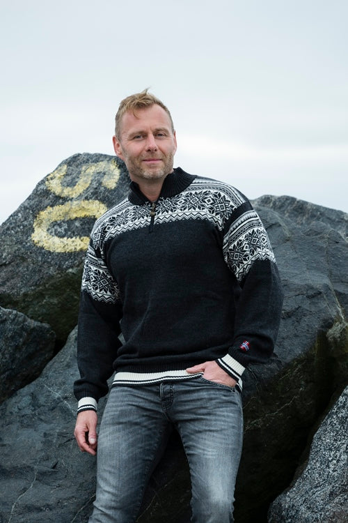 A person stands in front of large rocks, wearing the luxurious Arctic Circle Tor Merino Wool Sweater in grey and a pair of jeans.