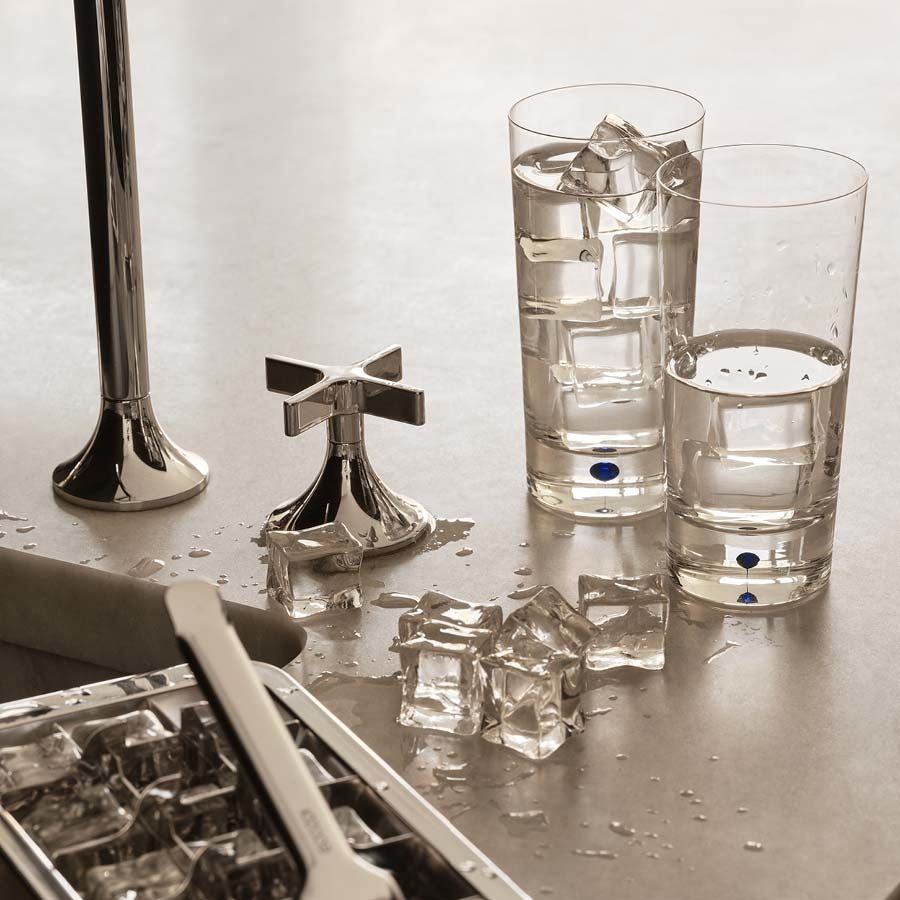 Two Orrefors: Intermezzo Blue Tumblers 13oz filled with ice and water sit on a countertop, with scattered ice cubes glistening near a chrome faucet and an ice tray.