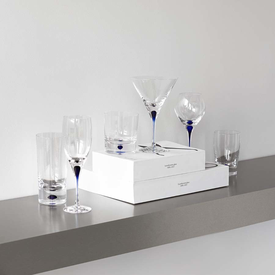 A display of exquisite Orrefors glassware by Erika Lagerbielke, including wine glasses and a martini glass with blue details, highlights the artistry of the Intermezzo Blue Tumbler design. The pieces are elegantly arranged on two white boxes on a shelf.