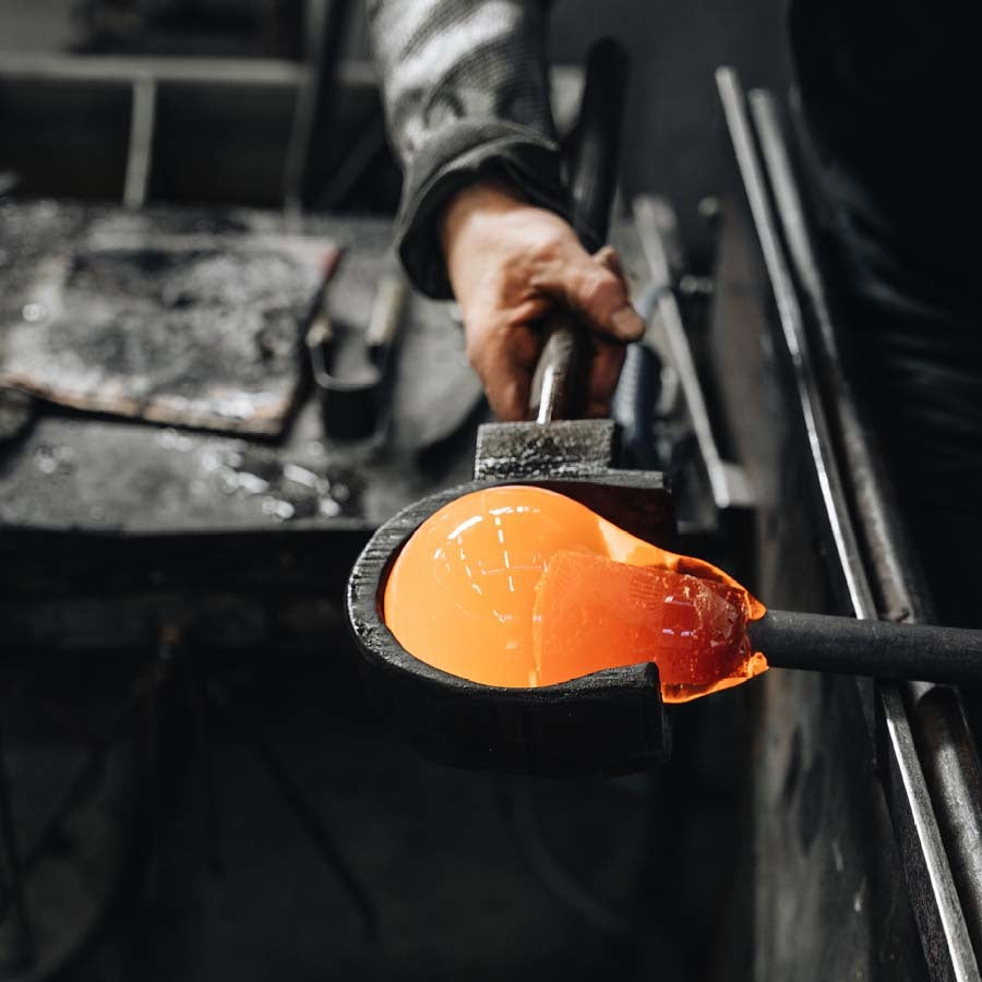 In a workshop, a craftsman shapes a glowing orange glass orb with metal tools, echoing the artistry of the Orrefors Intermezzo Blue Tumbler 13oz by Erika Lagerbielke.