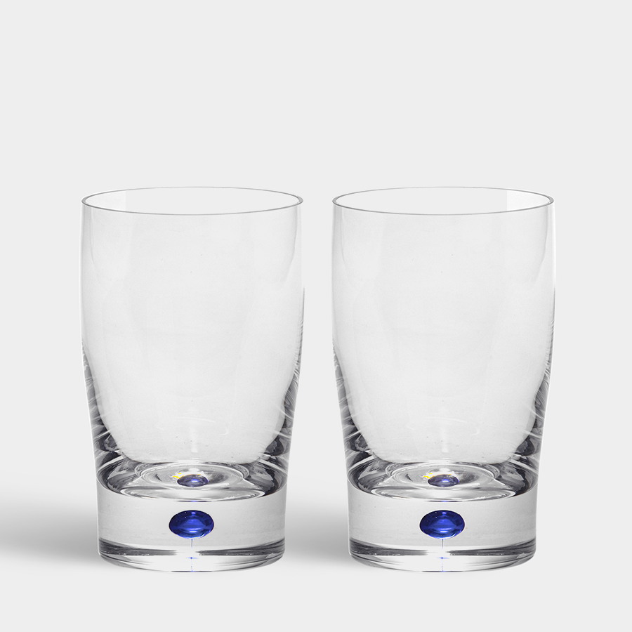 The Orrefors Intermezzo Blue Tumbler/Juice 9oz (2-Pack), designed by Erika Lagerbielke, features a small blue dot at the base. These clear glass tumblers elegantly stand against a white background, highlighting their simplicity.