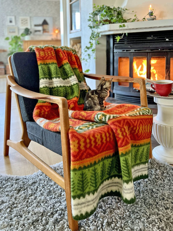 A cat sits on a chair draped with the Mora Blanket – 100% Norwegian Lamb’s Wool by Kerstin Landström Design near a lit fireplace, with a red cup on a small table in a cozy living room.