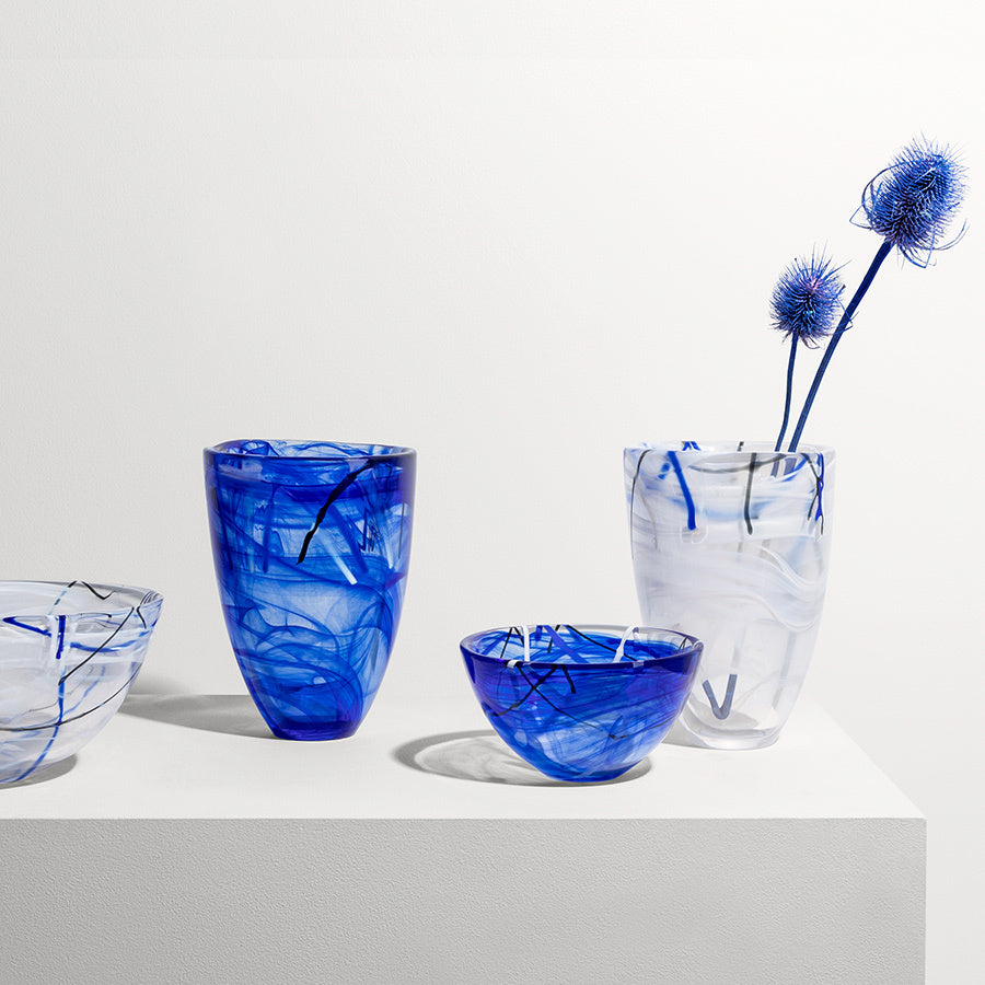 Four blue swirled glass vases and bowls from the KostaBoda: Contrast Vase Blue collection by Anna Ehrner, sit on a white surface; a tall clear vase elegantly holds three spiky blue flowers, beautifully contrasting bold blue hues with crystal clarity.