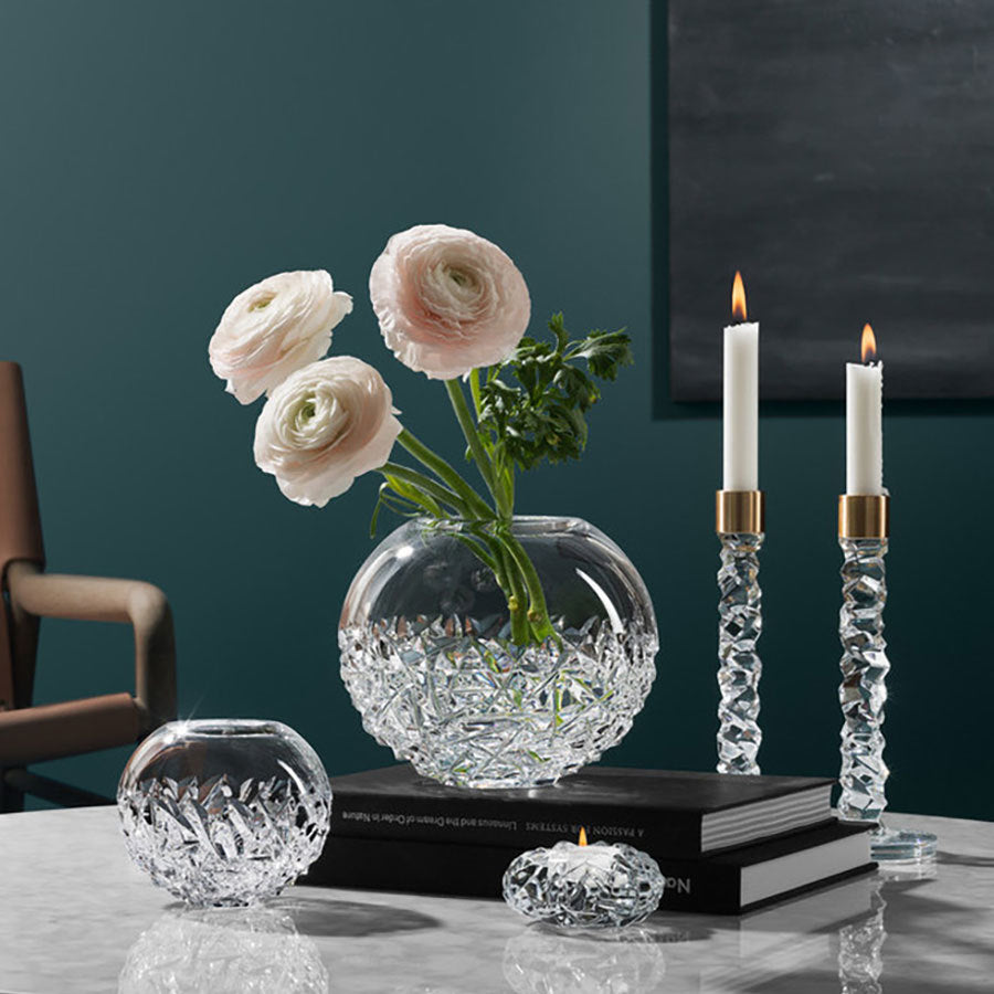 White flowers in an Orrefors: Carat Globe Vase Large are elegantly displayed next to crystal glass candle holders with lit candles on a marble surface with books. A dark green wall and part of a chair create a sophisticated backdrop, evoking an Orrefors display.