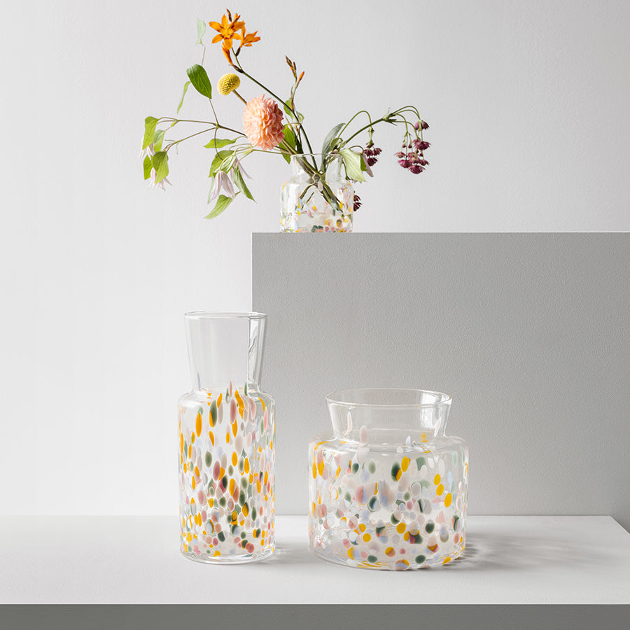 The KostaBoda: Meadow Vase Spring Tall, showcasing Sara Perssons artistry with its colorful dot patterns, stands proudly on an upper shelf filled with flowers, while two similar vases below await their blooms.