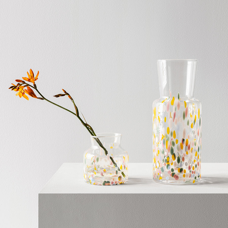 Two Kosta Boda glass vases rest on a white surface. The smaller Meadow Vase in Spring Tall by Sara Persson cradles an orange flower with a curved stem, set against a plain light gray wall.