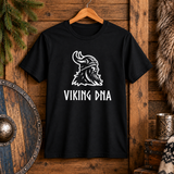 Black t-shirt with a white graphic of a bearded Viking wearing a horned helmet and the words “VIKING DNA,” displayed among rustic Viking heritage decor items.