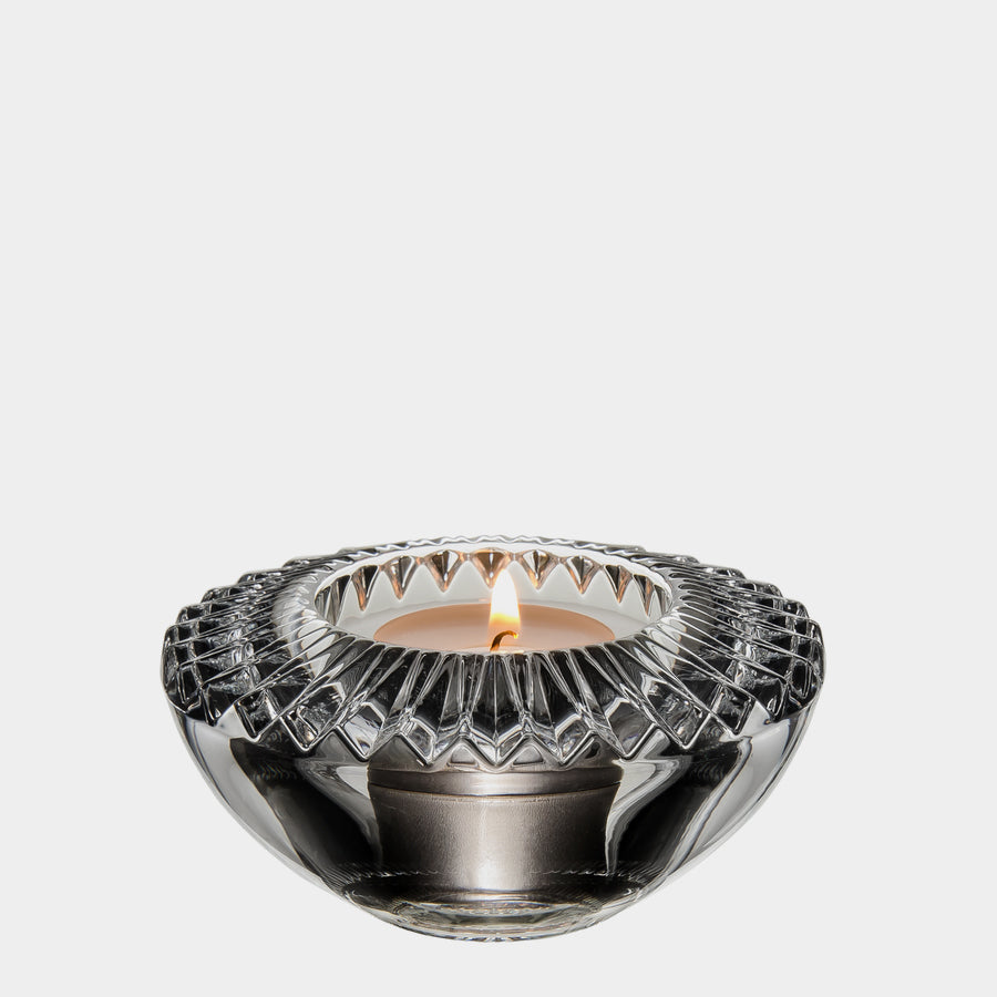 Orrefors Designs introduces the Brilliance Votive Candle Holder, a crystal glass piece featuring an intricate cut pattern that elegantly displays a lit votive candle against a plain white backdrop.