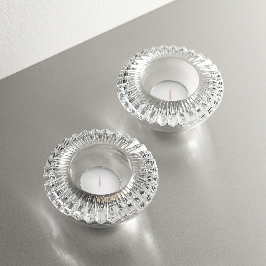 Two Orrefors: Brilliance Votive Candle Holders with tealight candles are set on a reflective surface, echoing their elegance.