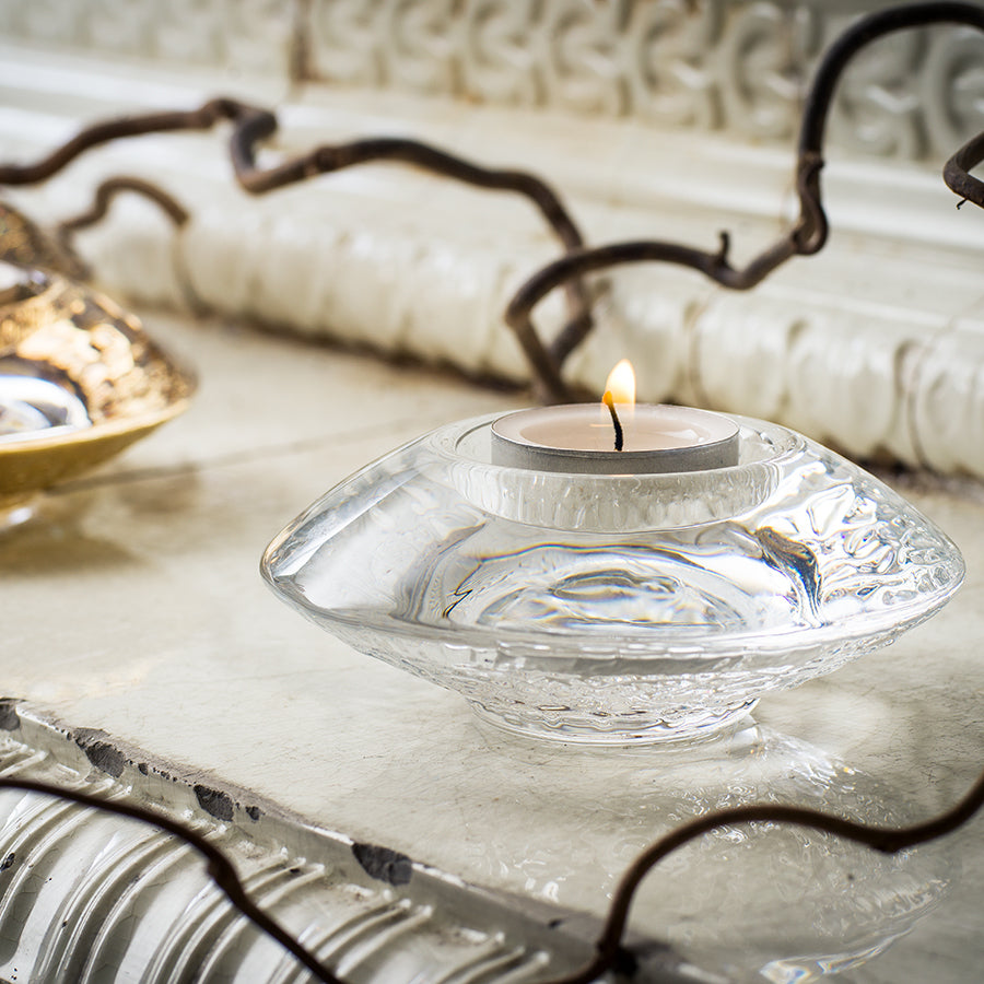 A lit tealight candle rests in an Orrefors Discus Clear Votive Small Candle Holder, designed by Lars Hellsten, shaped like a shallow discus and placed on an ornate white surface with decorative branches nearby.