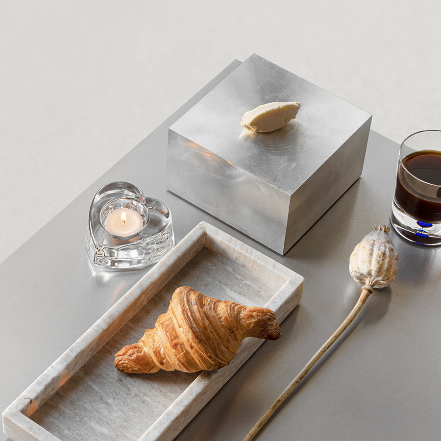 A croissant on a rectangular plate, an Orrefors My Heart Votive Candle Holder, a metal box with a butter pat, a dried poppy pod, and a glass of dark liquid elegantly arranged on a light gray surface.