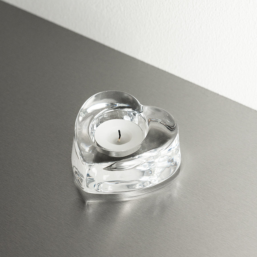 The Orrefors: My Heart Votive Candle Holder with a white tealight graces a gray surface, elegantly reflecting light and romance.