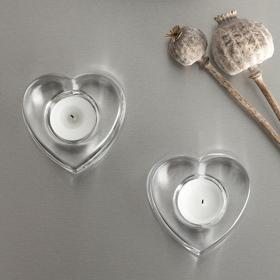 Two Orrefors: My Heart Votive Candle Holders, each with a white candle, sit on a gray surface beside dried poppy pods.