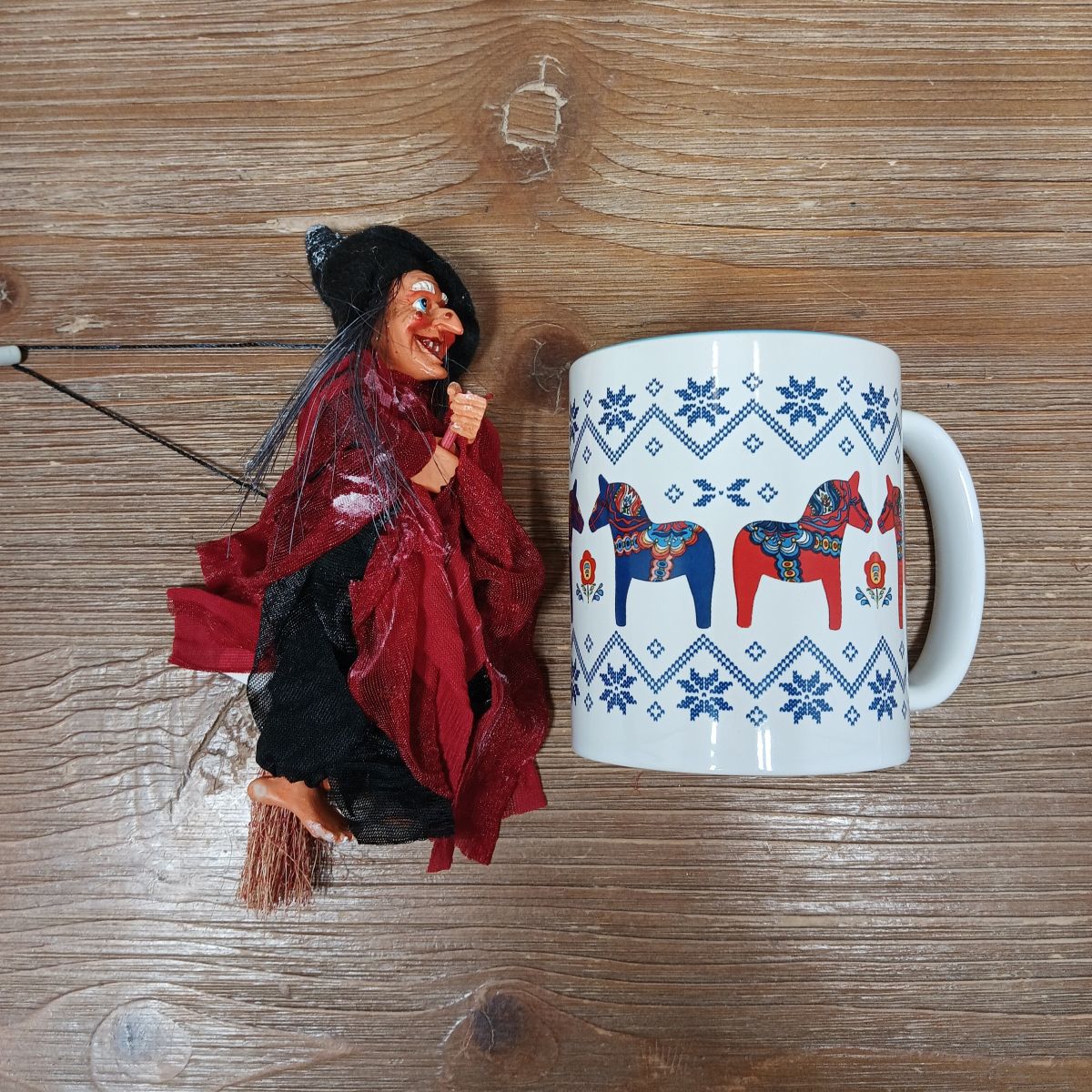 The Walburga Red Kitchen Witch Doll, handmade in Germany, stands next to a white mug featuring blue Swedish Dala horses and snowflake motifs on a wooden surface.
