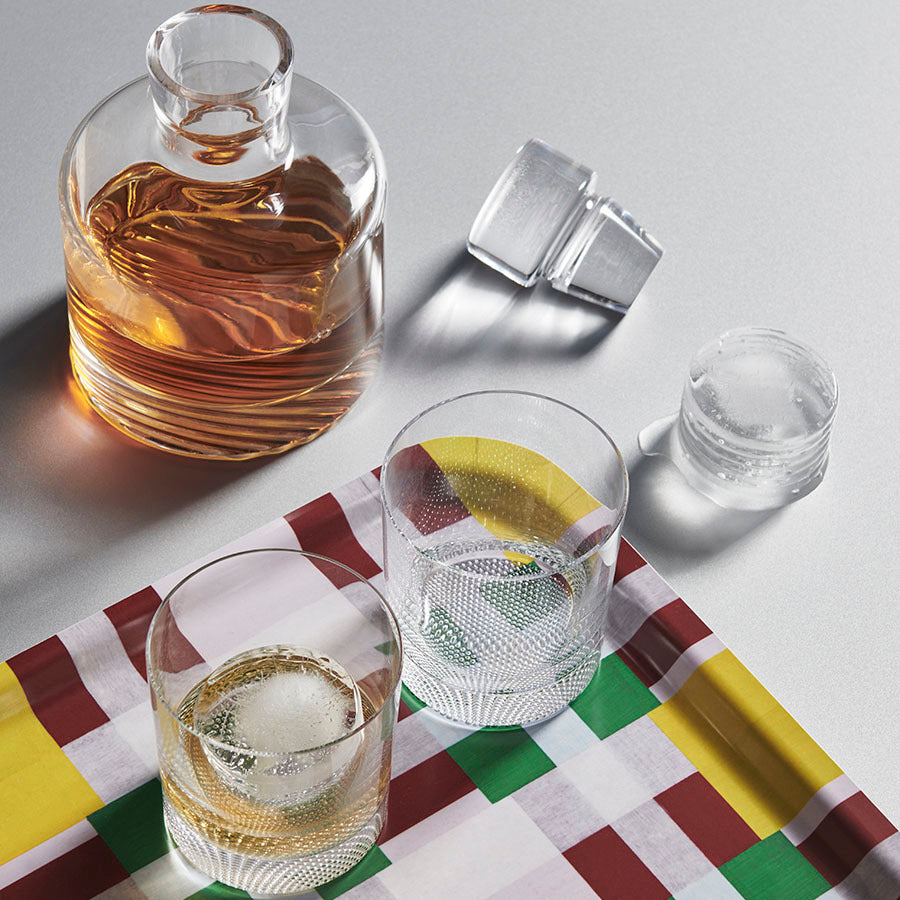 A clear glass decanter with amber liquid and two ice-filled glasses rest on a checkered cloth over a reflective surface, all part of the KostaBoda: Wave 3 Piece Gift Set, which includes 1 decanter and 2 double old fashioned glasses.