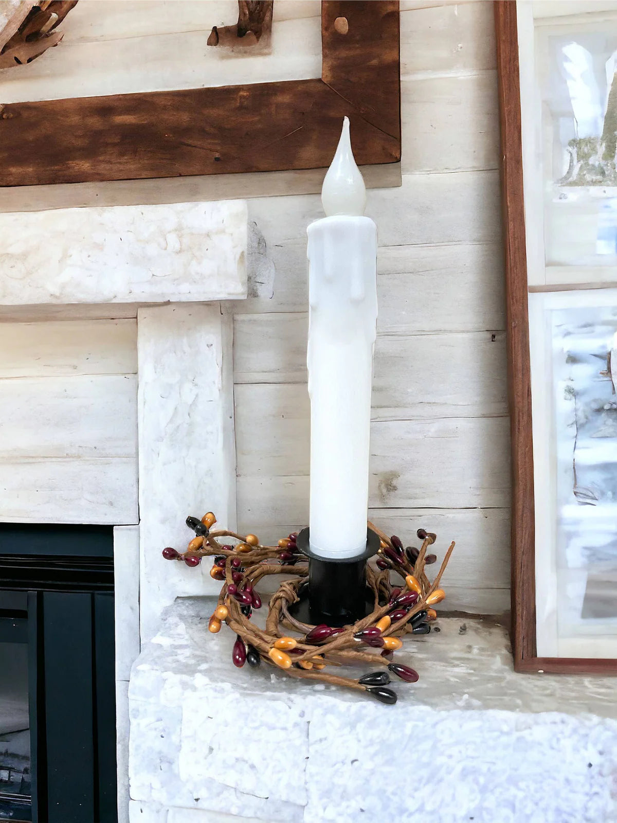 A Hand-Dipped Beeswax 7 Tapered LED Light, with a battery-operated timer feature, rests on the stone mantel amid a wreath of red and orange berries. It captures farmhouse charm alongside framed art, offering a cozy and elegant ambiance.