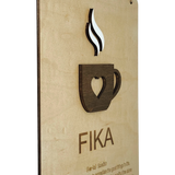 Wooden plaque featuring a heart-shaped coffee cup cutout with rising steam and the word “FIKA” below, displayed on a light wood background.