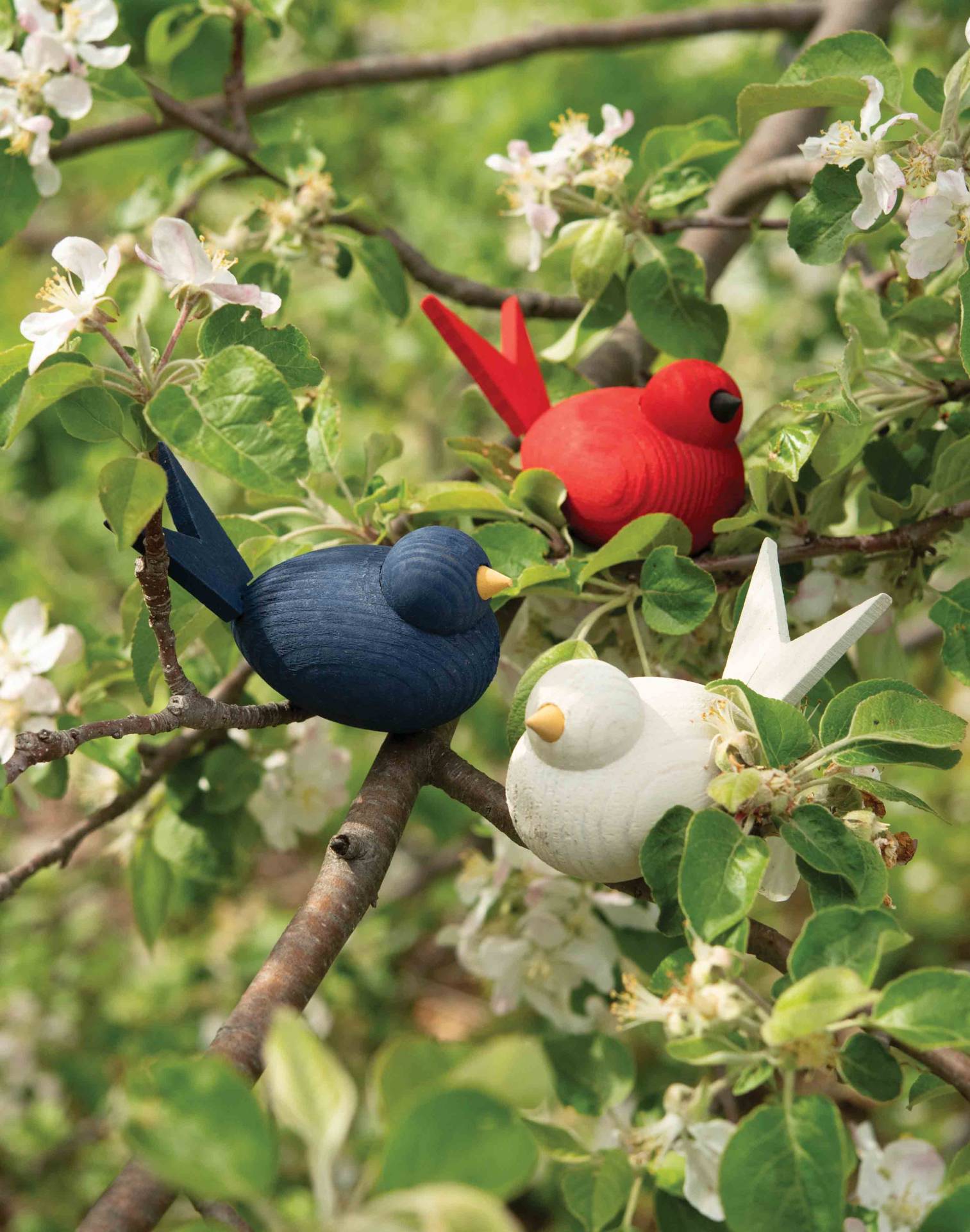 Three vibrant handcrafted Swedish wooden bird figurines in red, white, and blue are perched on branches adorned with green leaves and white blossoms.