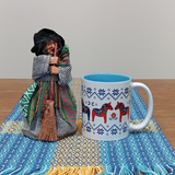 A Yasmina Green Kitchen Witch Doll figurine (7" tall) with a standing base is placed beside a white mug featuring red and blue horses, both set on a woven blue-and-yellow cloth atop a wooden table.