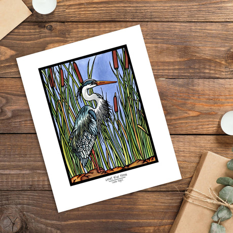 The Artwork: Blue Heron 8x10 Print by Sarah Angst is a giclee print featuring a great blue heron among tall reeds, displayed on wood beside a candle, envelope, and wrapped gift.